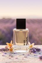 A clear rectangular perfume bottle with a black cap stands on a stone surface surrounded by lavender flowers, with a blurred lavender field in the background. The bottle label reads NUR FLORAL.